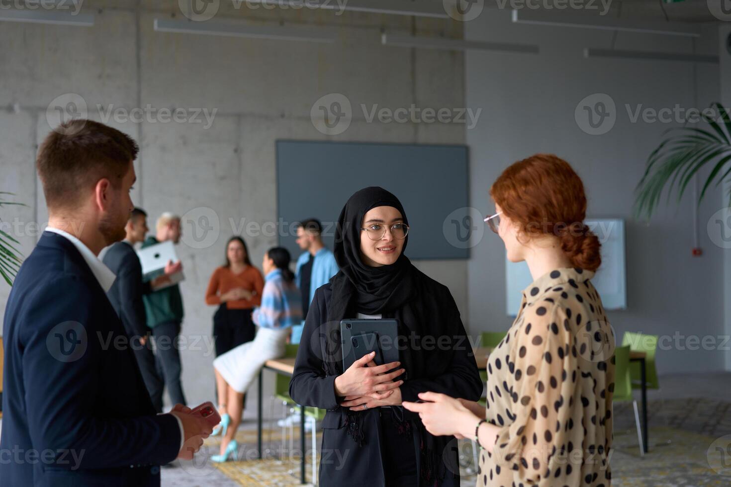 A diverse group of business professionals engages in a conversation during a work break in the office, with their colleagues visible in the background, fostering collaboration and connection photo