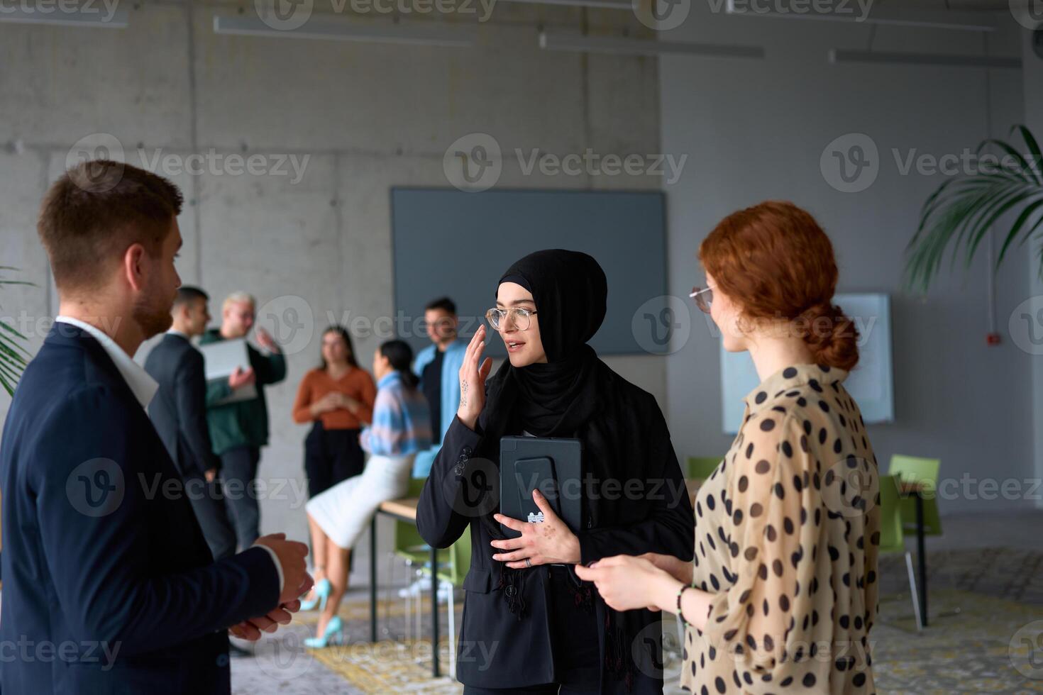 A diverse group of business professionals engages in a conversation during a work break in the office, with their colleagues visible in the background, fostering collaboration and connection photo