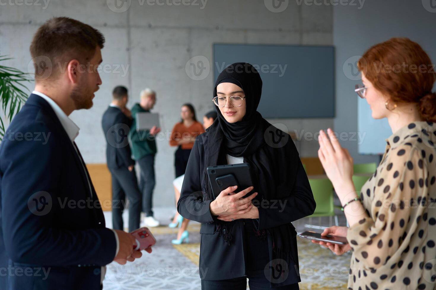 A diverse group of business professionals engages in a conversation during a work break in the office, with their colleagues visible in the background, fostering collaboration and connection photo