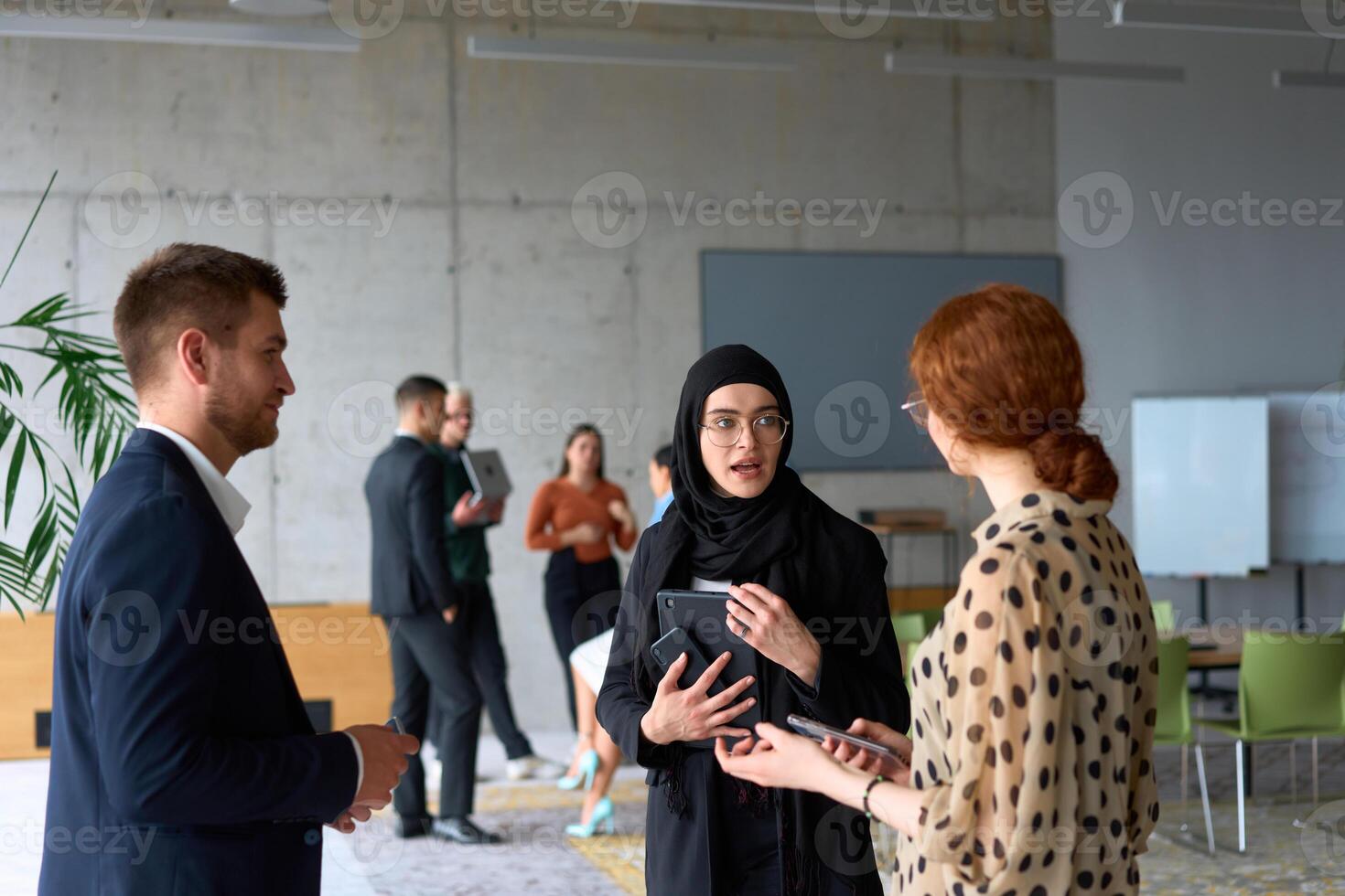 A diverse group of business professionals engages in a conversation during a work break in the office, with their colleagues visible in the background, fostering collaboration and connection photo