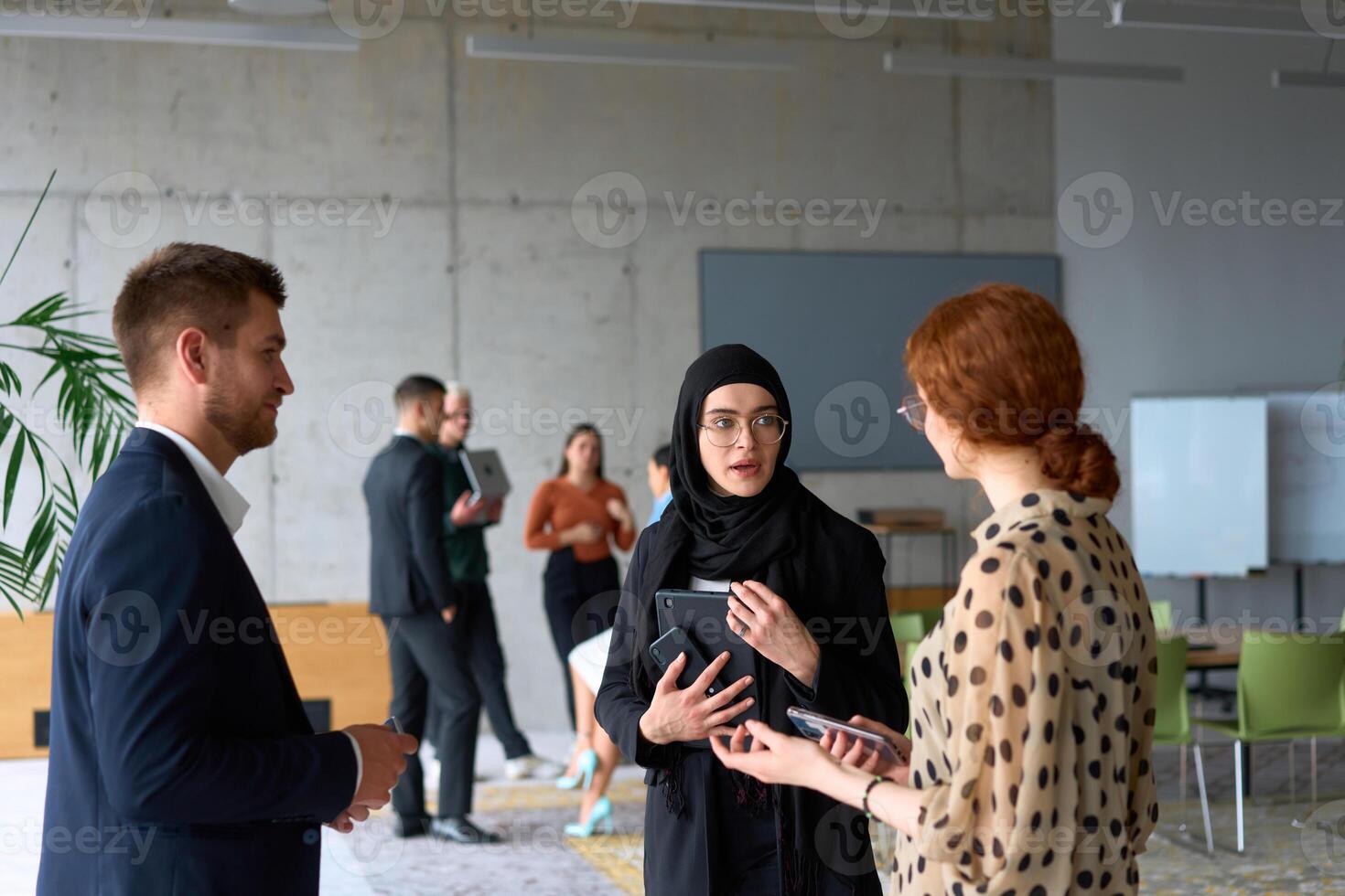 A diverse group of business professionals engages in a conversation during a work break in the office, with their colleagues visible in the background, fostering collaboration and connection photo