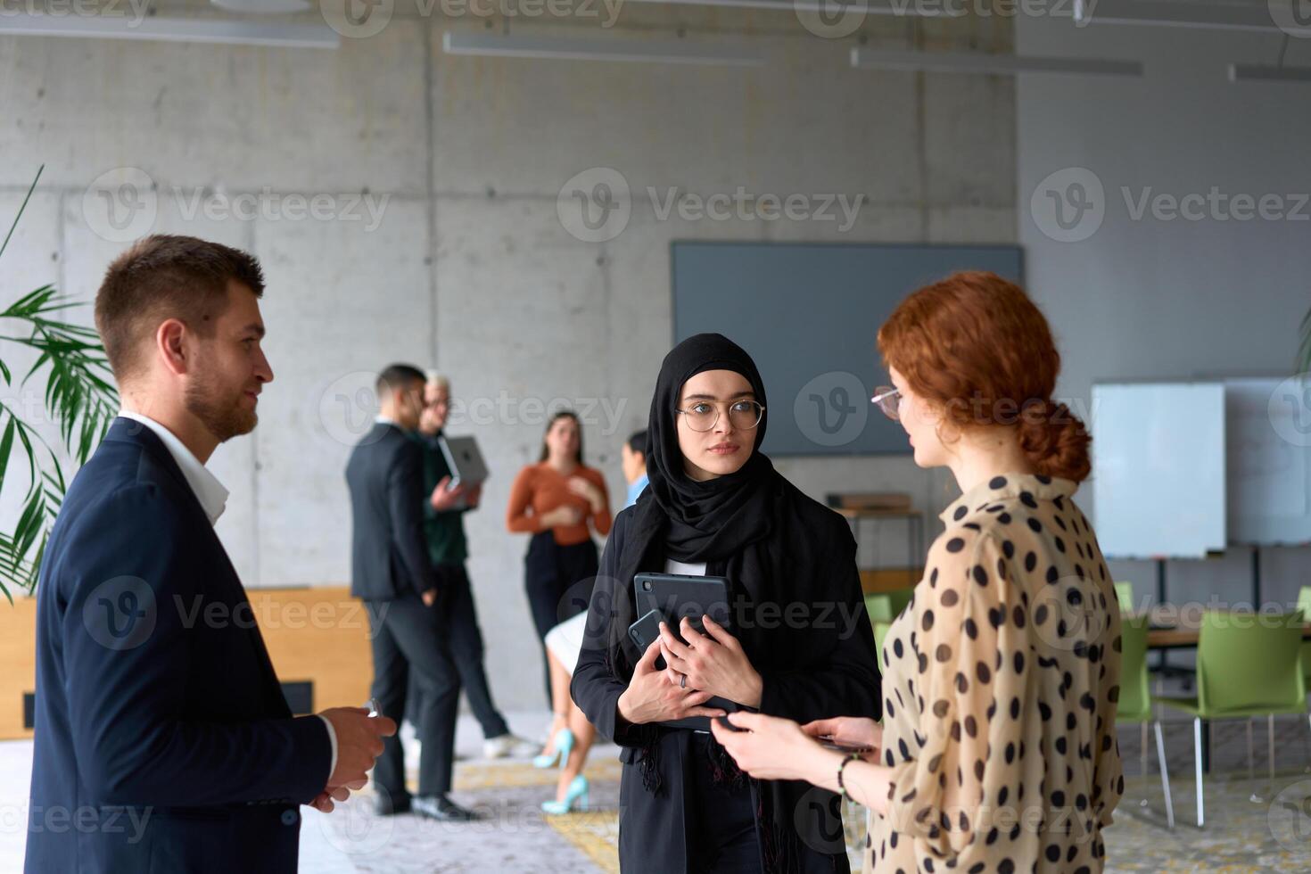 A diverse group of business professionals engages in a conversation during a work break in the office, with their colleagues visible in the background, fostering collaboration and connection photo