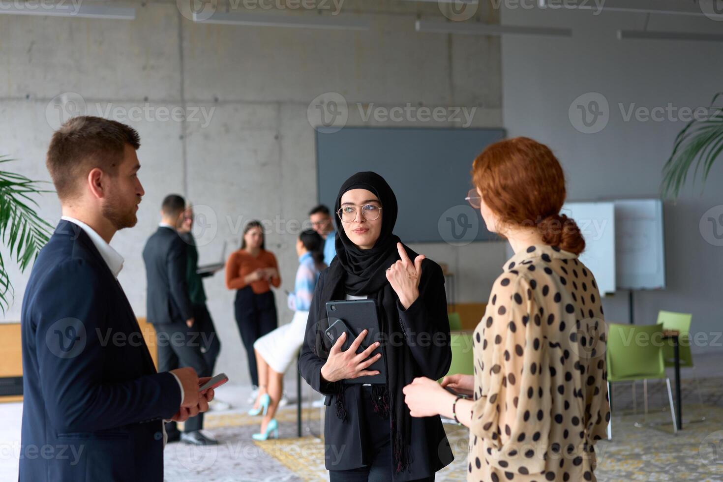 A diverse group of business professionals engages in a conversation during a work break in the office, with their colleagues visible in the background, fostering collaboration and connection photo