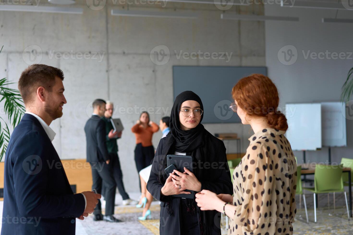 A diverse group of business professionals engages in a conversation during a work break in the office, with their colleagues visible in the background, fostering collaboration and connection photo