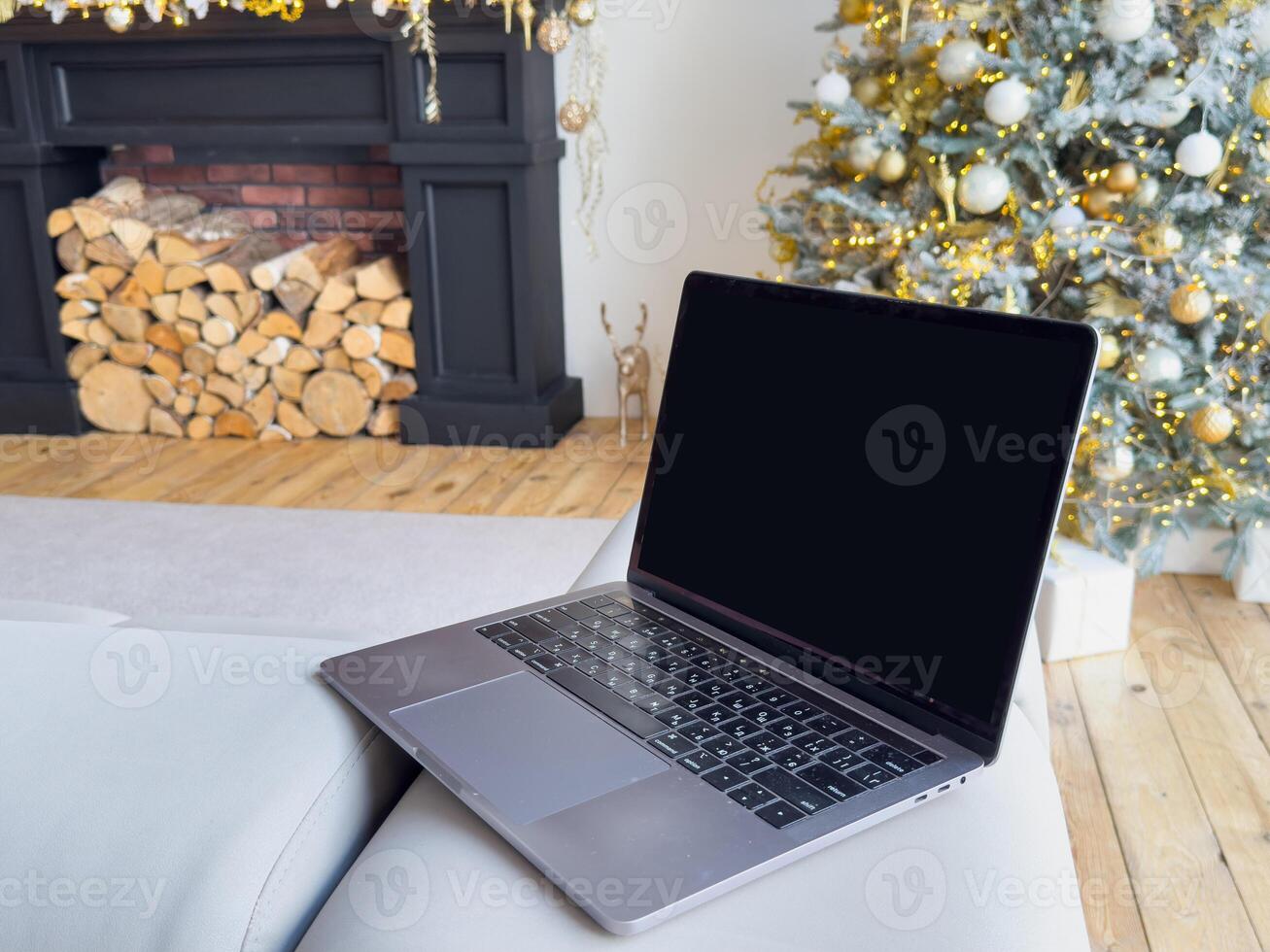 Laptop with empty screen and stylish christmas gifts on background of fireplace, tree with golden lights in festive cozy room. Christmas shopping online and sales. Laptop mock up, copy space photo