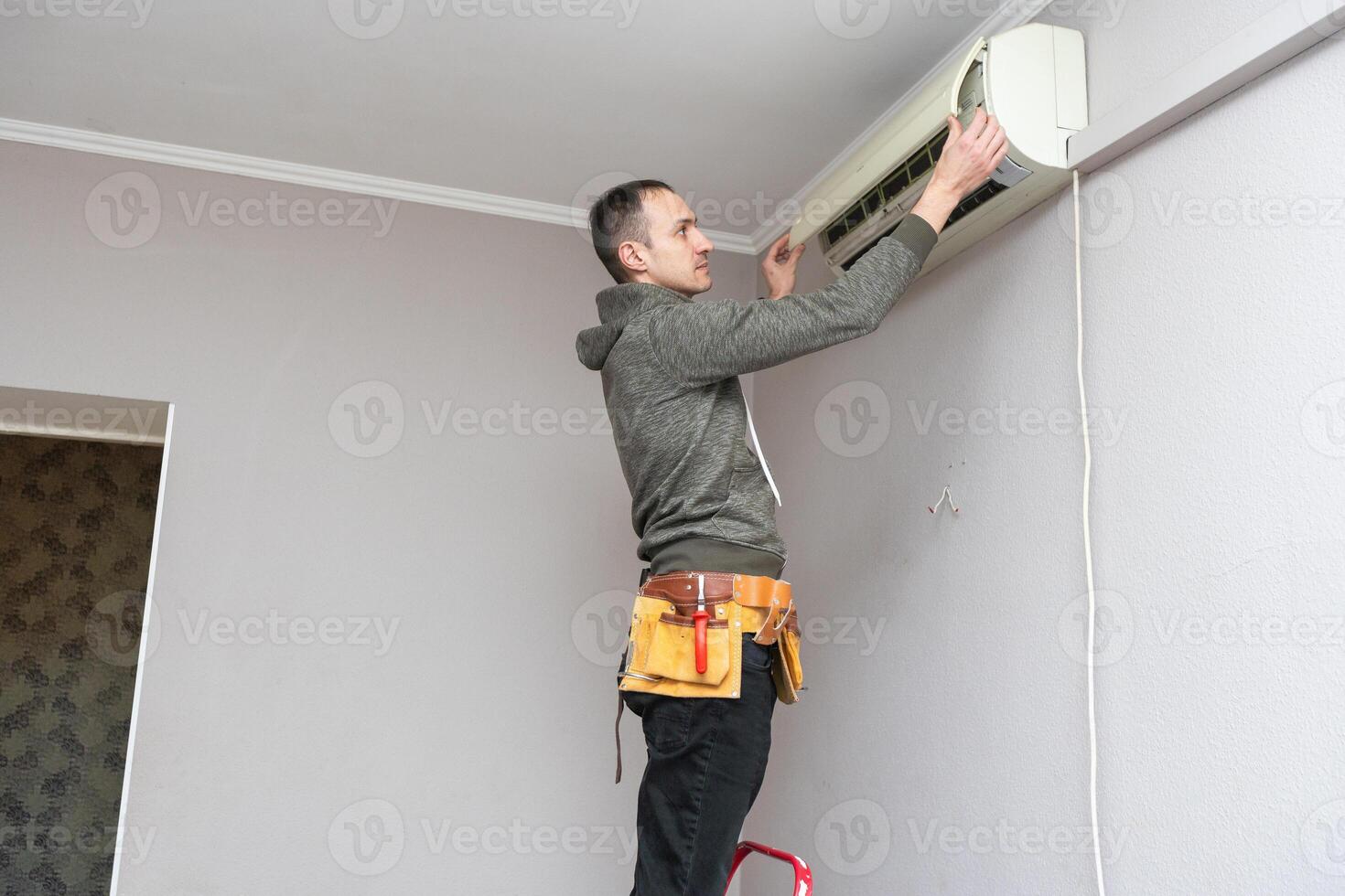 Professional electrician man with screwdriver maintaining, cleaning modern air conditioner indoors. Young technician standing on a ladder in the room repairing or installing air conditioner. photo
