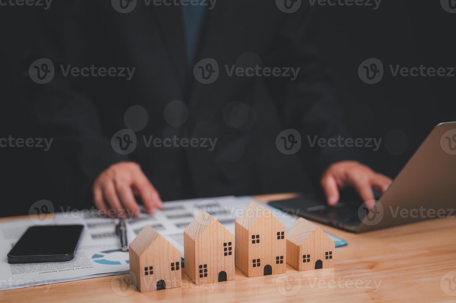 Professional evaluating real estate investments using wooden house models, laptop, and documents image captures essence of strategic decision-making in property investment photo