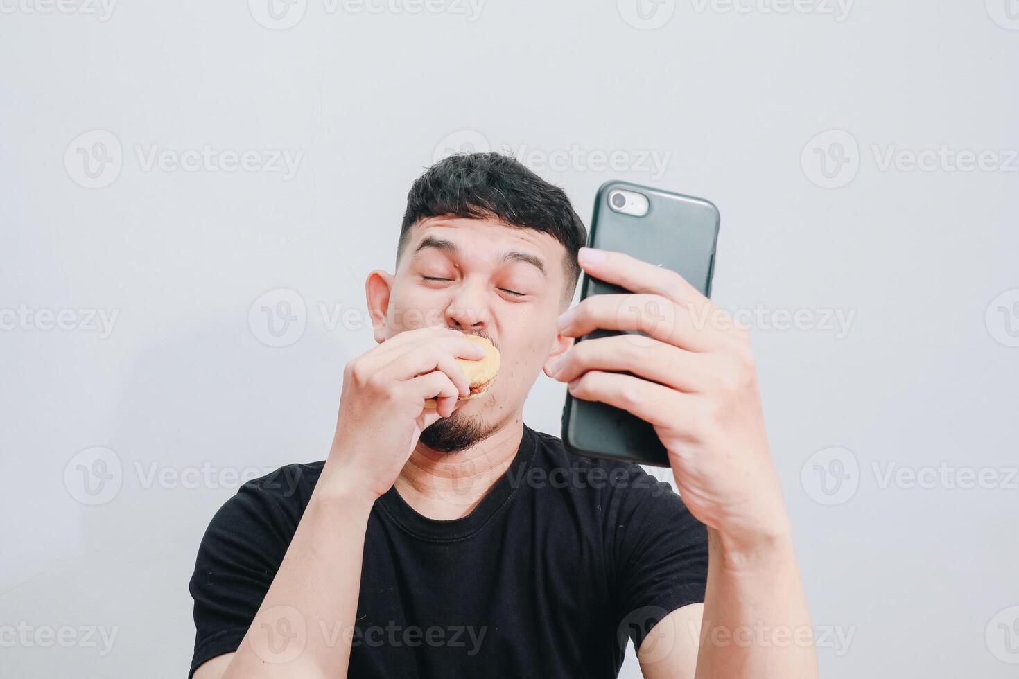 an asian man is enjoying his hamburger while holding his smartphone photo