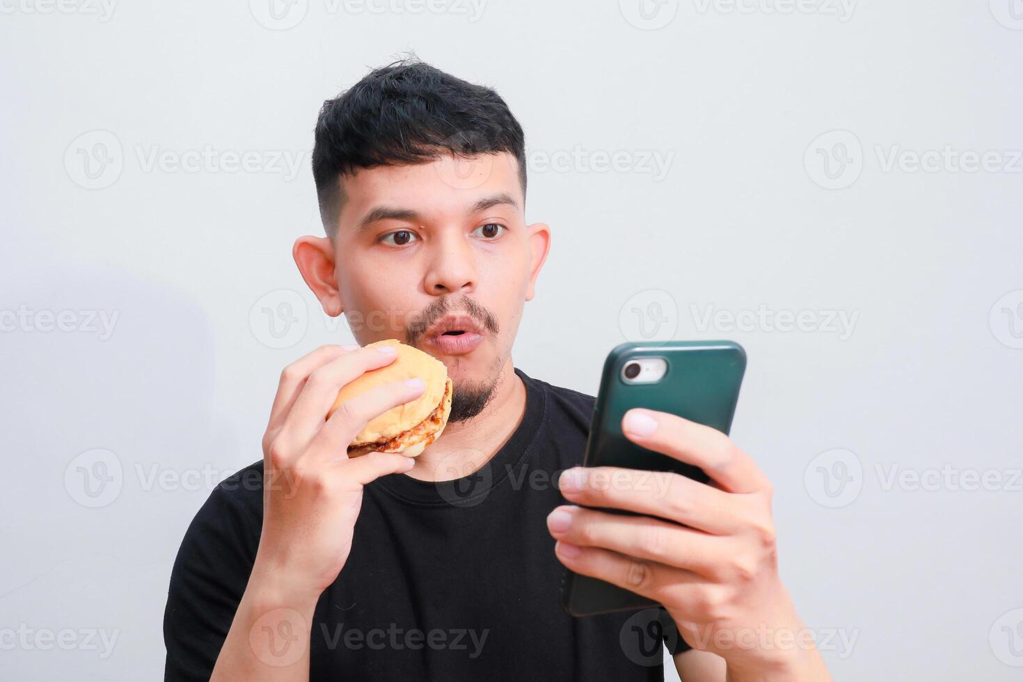 an asian man is enjoying his hamburger while holding his smartphone photo