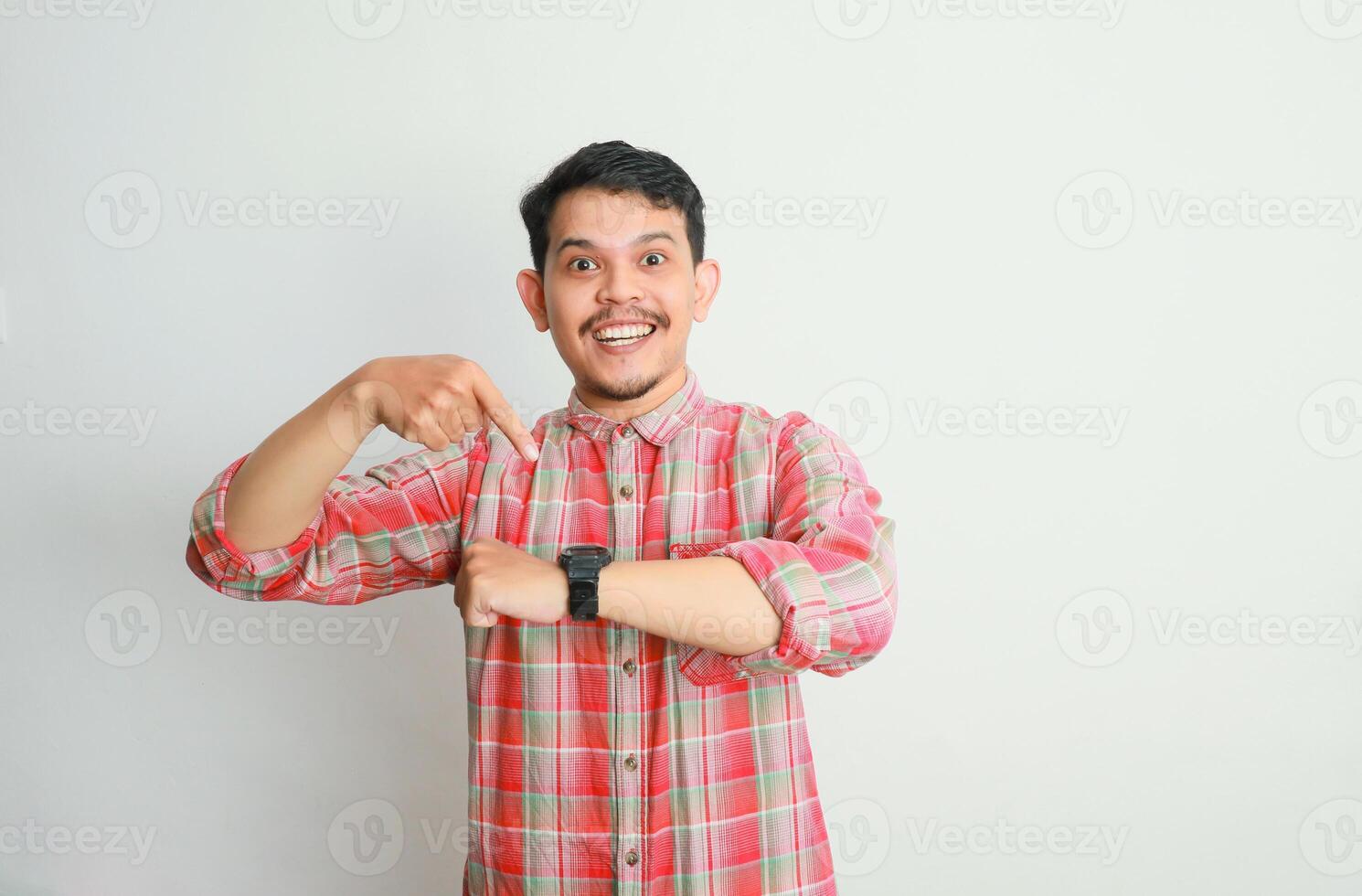 Portrait of excited Asian man in casual shirt looking at his wrist watch, pointing on it to remind the time. Advertising concept. Isolated image on white background photo
