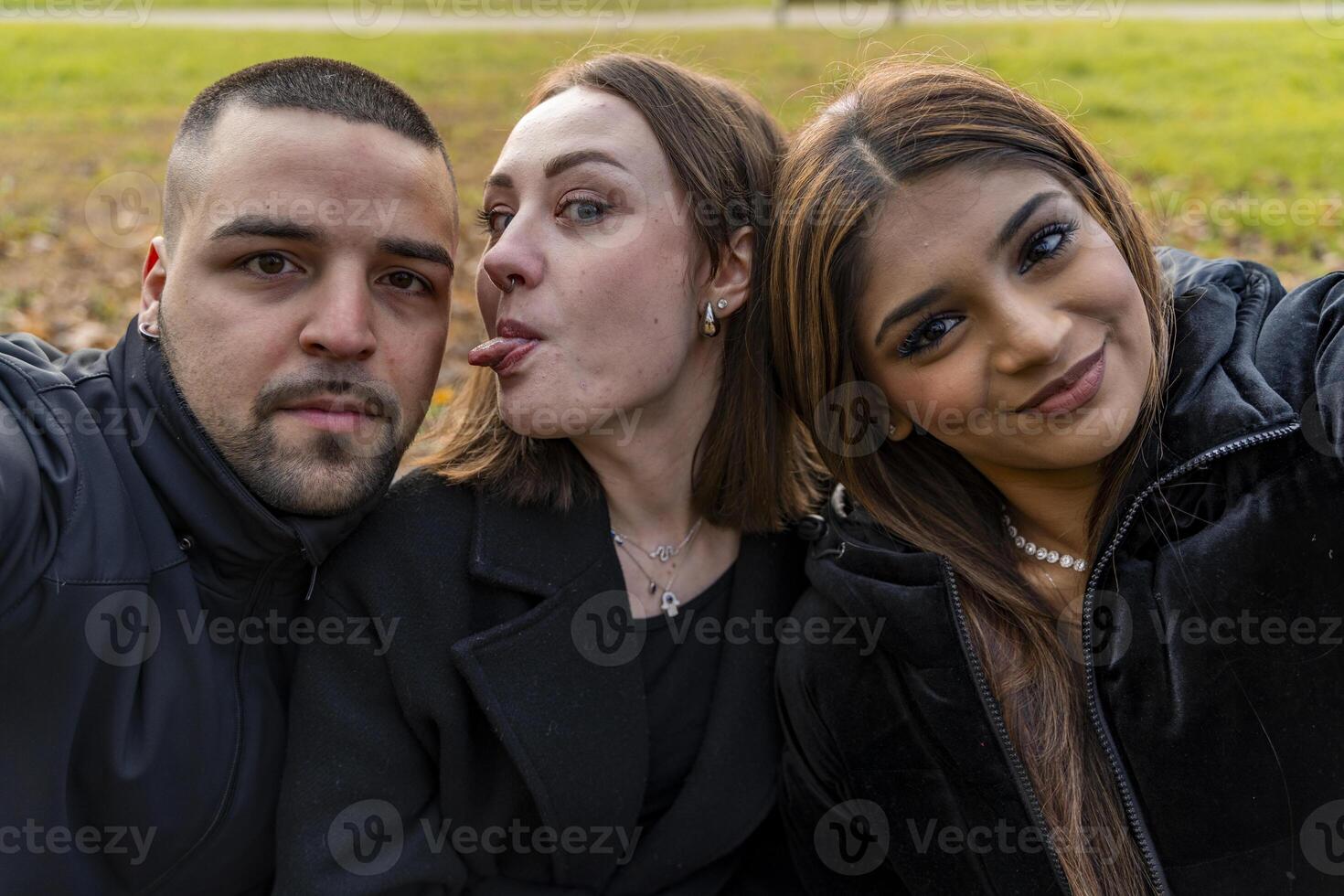 three young male and female friends take a selfie having fun sitting on a bench in a public park in winter photo