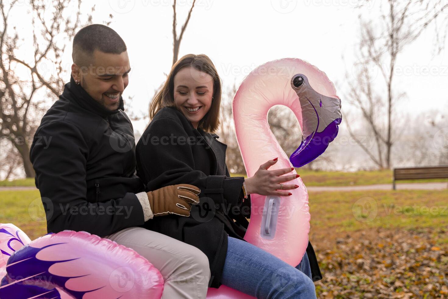 portrait of two young lovers having fun riding an inflatable pink flamingo in a public park in winter photo