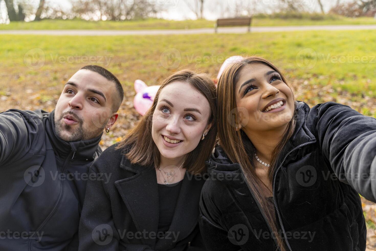 three young male and female friends take a selfie having fun sitting on a bench in a public park in winter photo