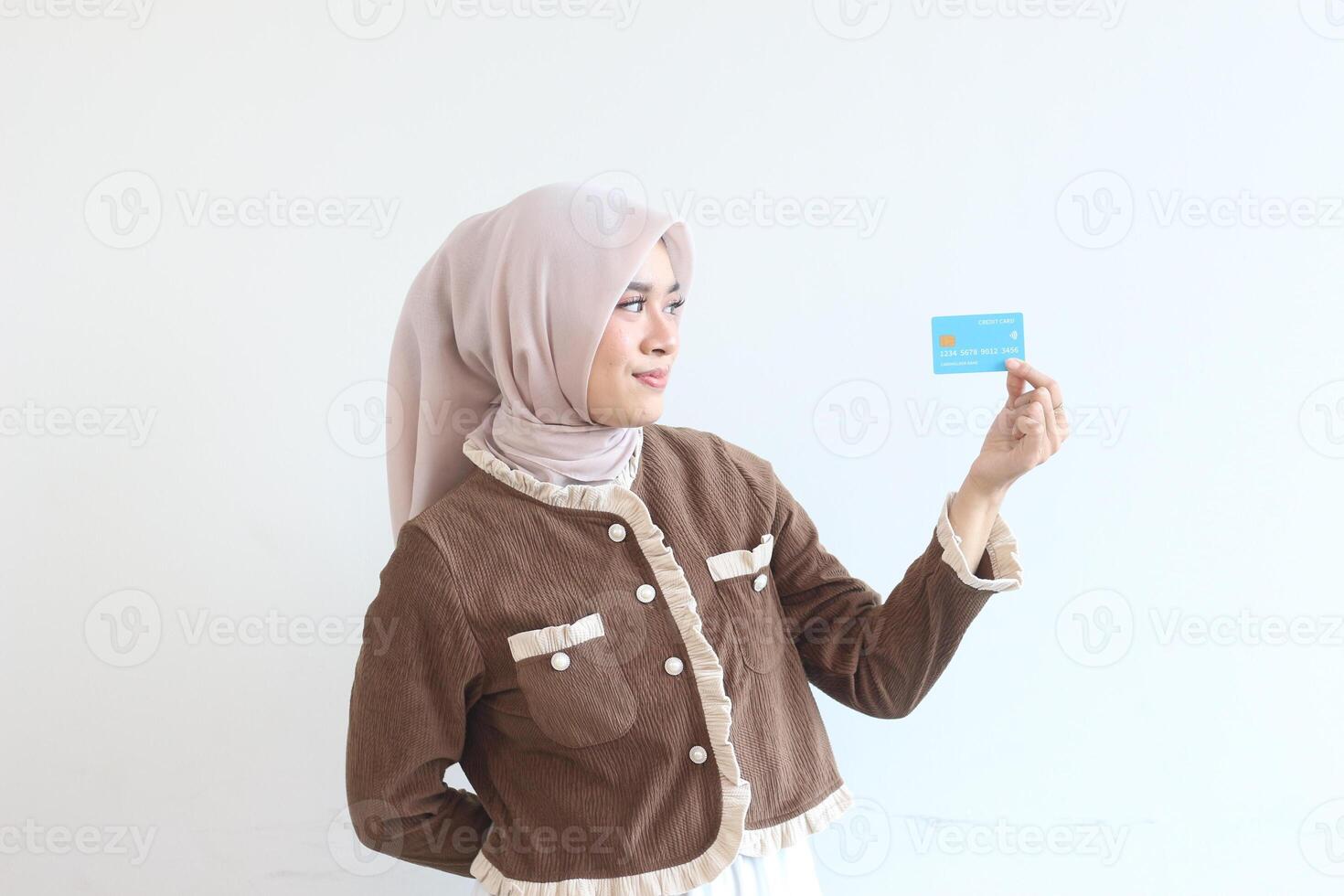 A Young Woman in a Brown Jacket and Beige Hijab Holding a Light Blue Credit Card Against a White Background photo