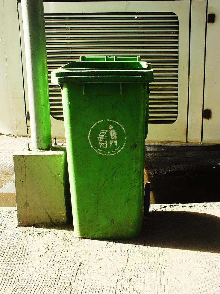 A green trash can with a sticker on it, sits empty on the ground. The trash can is located next to a metal pole and a building photo
