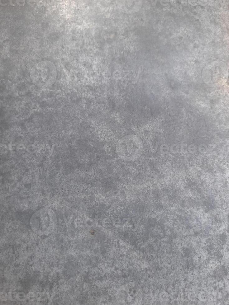 a close-up view of a textured surface a metal sheet, with a dull grayish tone. The surface features a mix of light and dark patches, creating a mottled, weathered, or oxidized appearance photo