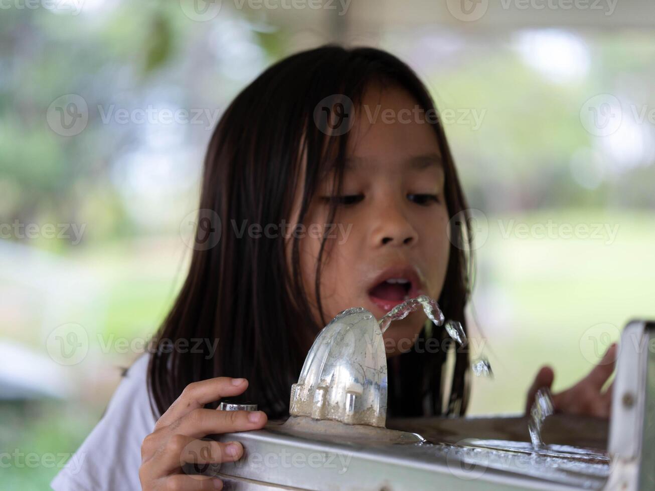 Cute girl drinks water from square shiny metal drinking fountain in a park. Children drink water from an outdoor public water dispenser. photo