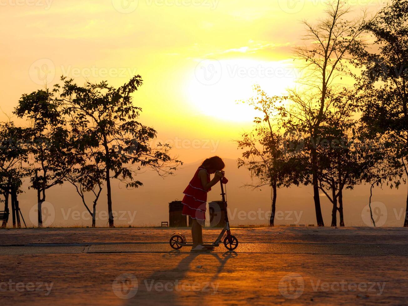Silhouette of a cute little girl riding a scooter in a park during a summer sunset at a public park. Active leisure and outdoor sport for child. photo