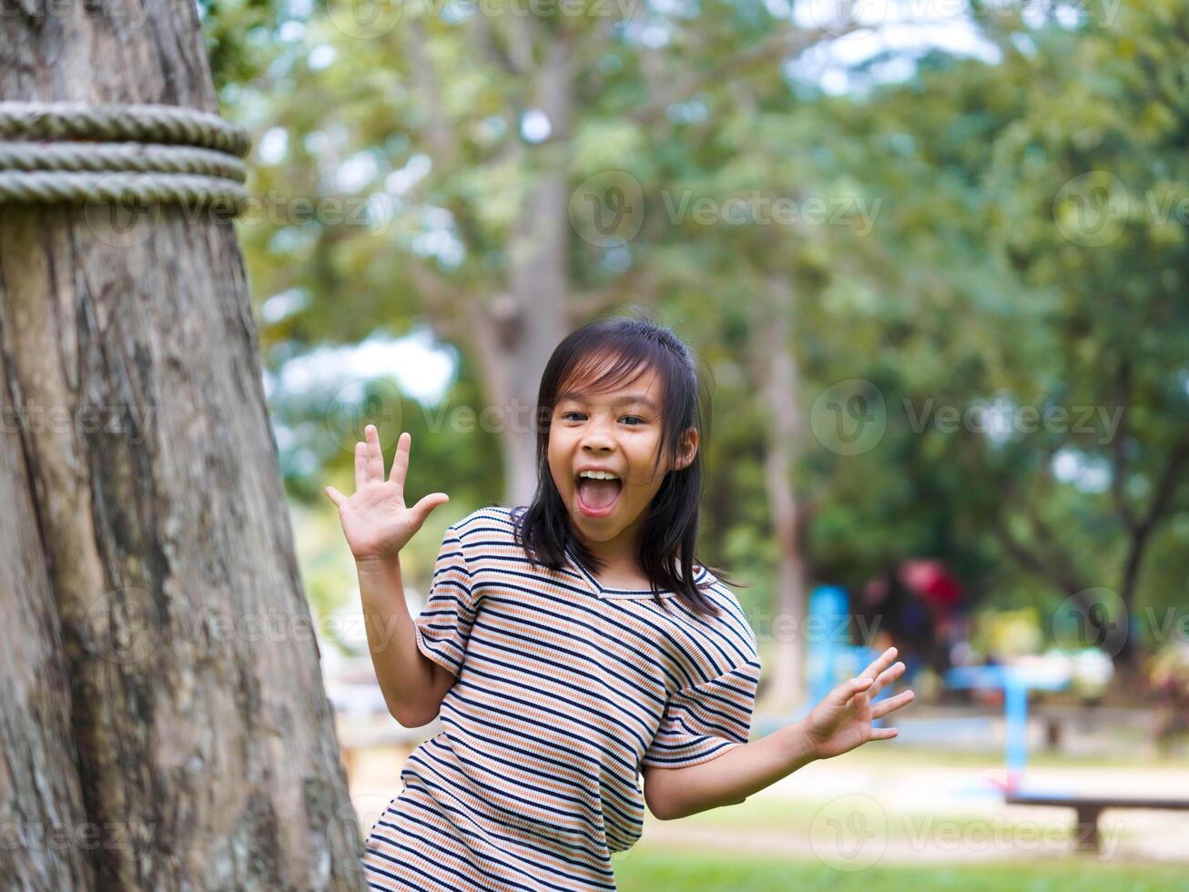 Cute little girl is peeking behind a tree, having fun playing playing peek-a-boo in a sunny garden. Smiling child in the park photo