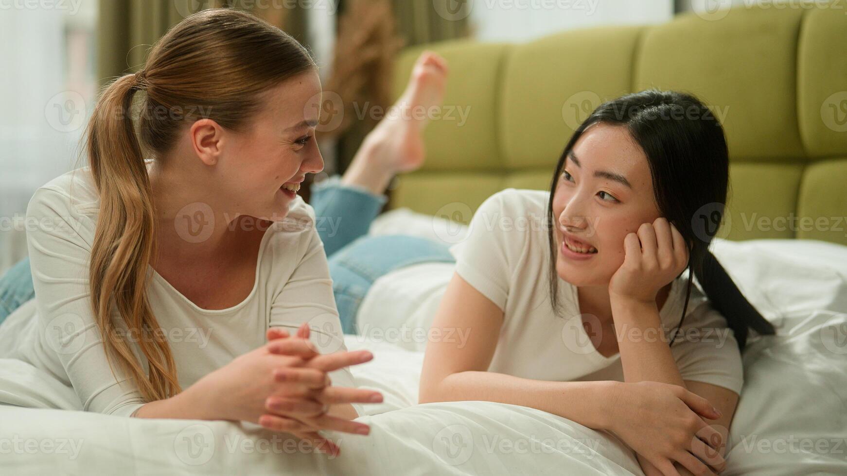 Two happy multiracial multiethnic women friends female diversity girls Caucasian woman talking with Asian girl share news laughing smiling showing thumb up applause gossiping on bed bedroom friendship photo