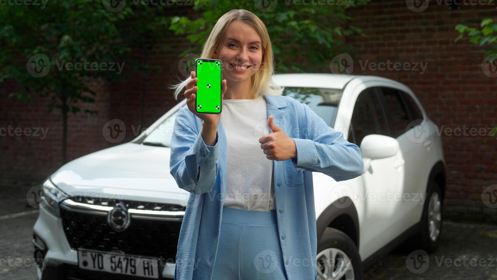 Smiling Caucasian girl female woman holding mobile phone tech green screen app chromakey automobile car driver showing like thumb up in city happy businesswoman client satisfied with auto rent service photo