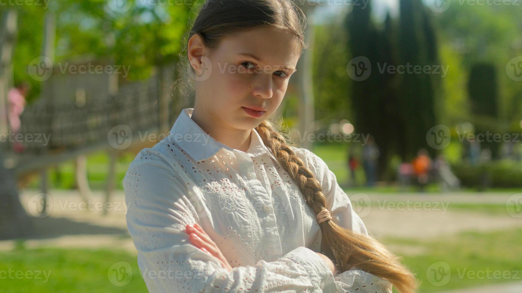 Upset sad frown little kid girl serious child daughter pupil female offspring in city portrait schoolgirl posing crossed hands offense family custody worried teenager alone in park playground outdoors photo
