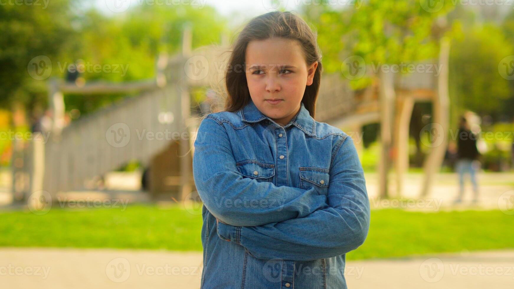 Portrait in park city outdoors offended angry alone sad serious Caucasian girl little child kid daughter orphan adopted custody family bad mood frowning schoolgirl pupil posing crossed hands offense photo