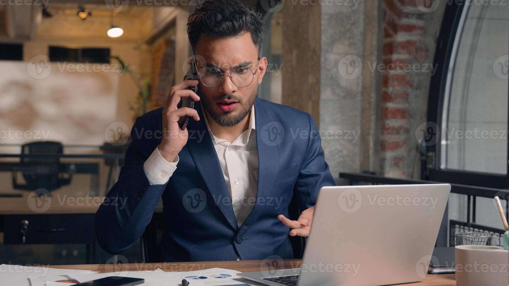 Stressed multitasking businessman Indian Arabian male guy annoyed employer manager consultant talking mobile phone shout working laptop writing documents anxious business man problem overwork office photo