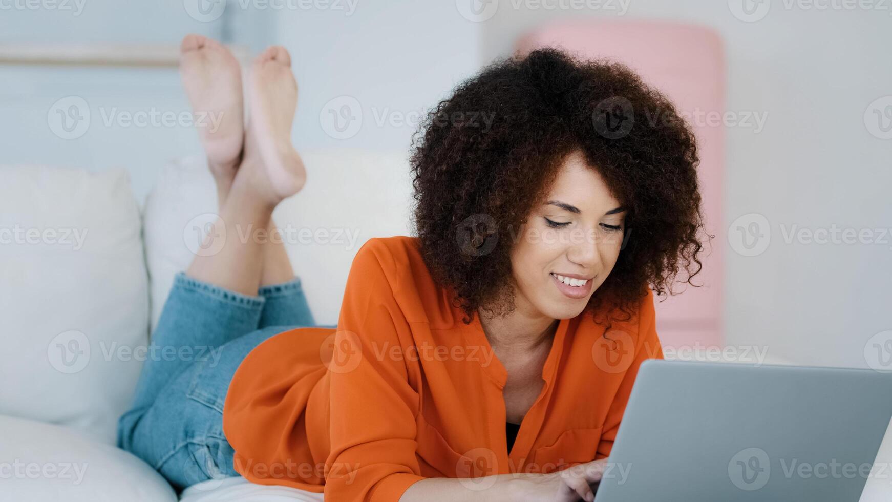 African 20s lady 30s woman freelancer with curly hair lying on sofa relaxing typing on laptop making online purchase in internet store app working remotely from home chatting with friends with laptop photo