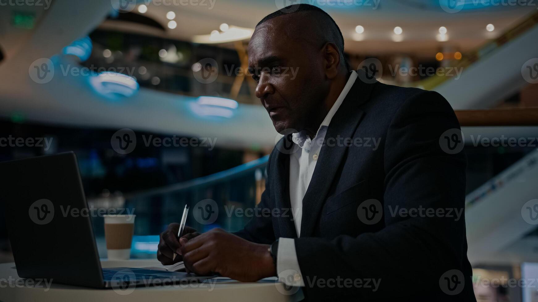 African middle-aged adult businessman worker entrepreneur man boss leader specialist company CEO freelancer teacher working with laptop writing notes in notebook check data in computer write in cafe photo