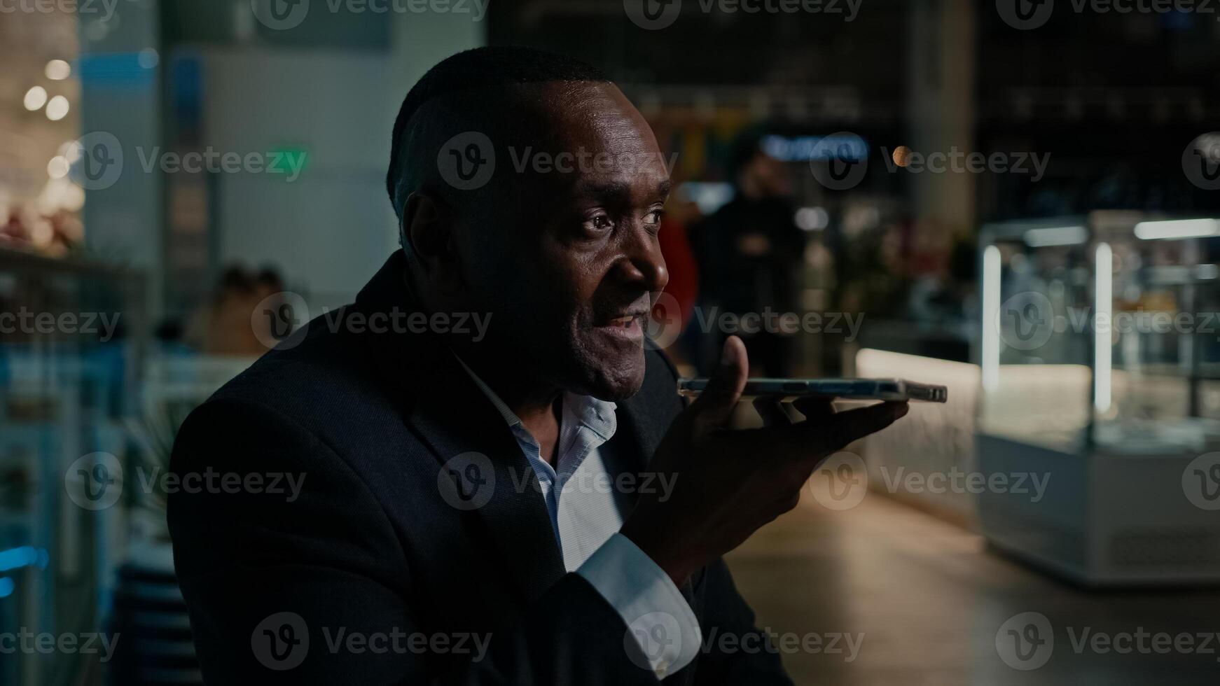African American senior mature middle-aged man recording voice recognition message on speakerphone. 50s ethnic businessman using virtual assistant phone app sets reminder on smartphone at mall cafe photo