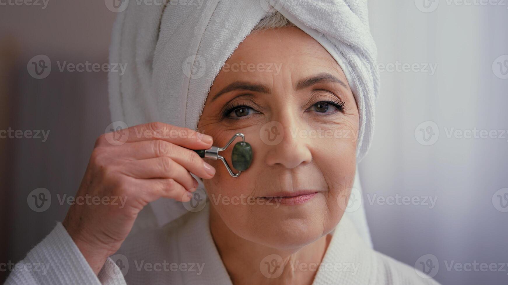 60s middle-aged old senior mature Caucasian woman with towel on head in bath morning face massage with stone roller elderly beautiful lady skincare procedure facial yoga self care exercise for skin photo