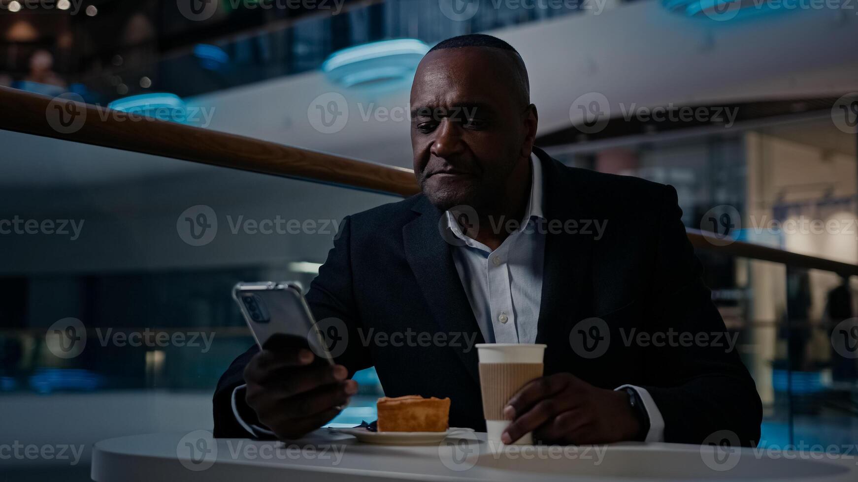 African adult middle-aged businessman 50s ethnic man in formal suit employer entrepreneur sitting at table in cafe drink coffee drinking tea browsing mobile phone smartphone app talk tell call photo