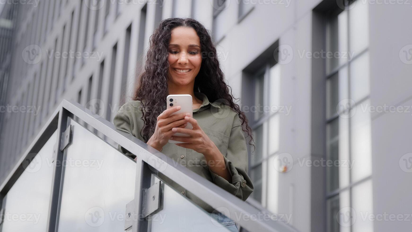 Smiling Caucasian Hispanic happy satisfied woman user lady standing un city outdoors on stairs chatting texting searching in net with mobile app phone browsing smartphone friendly communication online photo