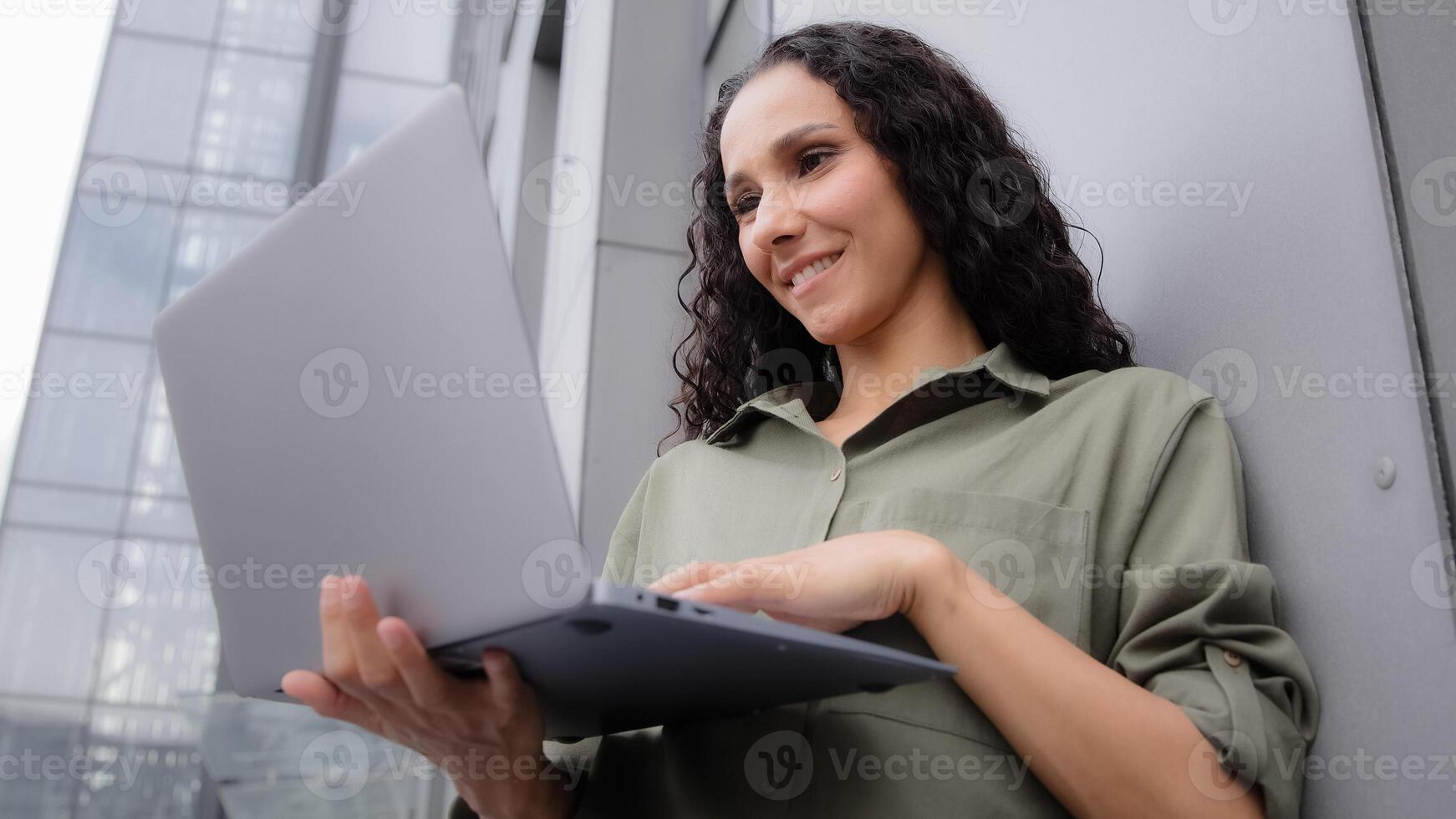 Close up busy smiling trader architect businesswoman boss user female investor lady leaning wall outdoors near company building working typing on laptop checking internet project on wireless computer photo