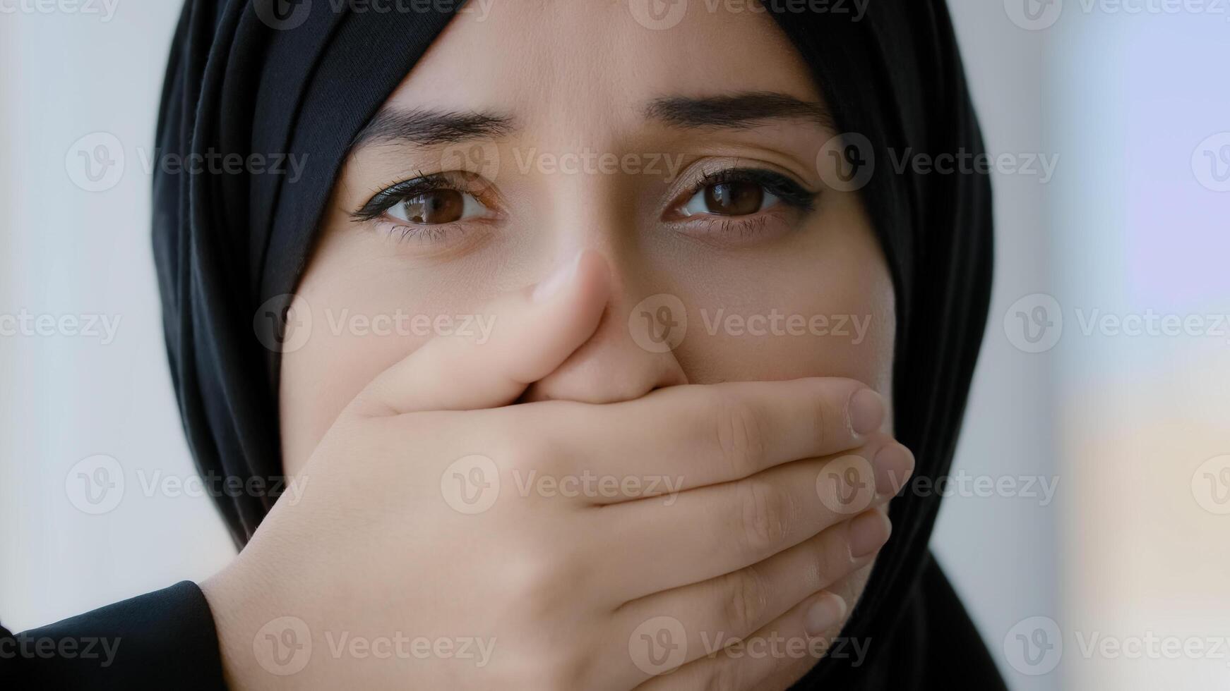 Female portrait indoors Islamic Arabian girl sad afraid Muslim woman Arab lady in black hijab looking at camera covers mouth with hands quiet silence discrimination stop talking oppression of women photo