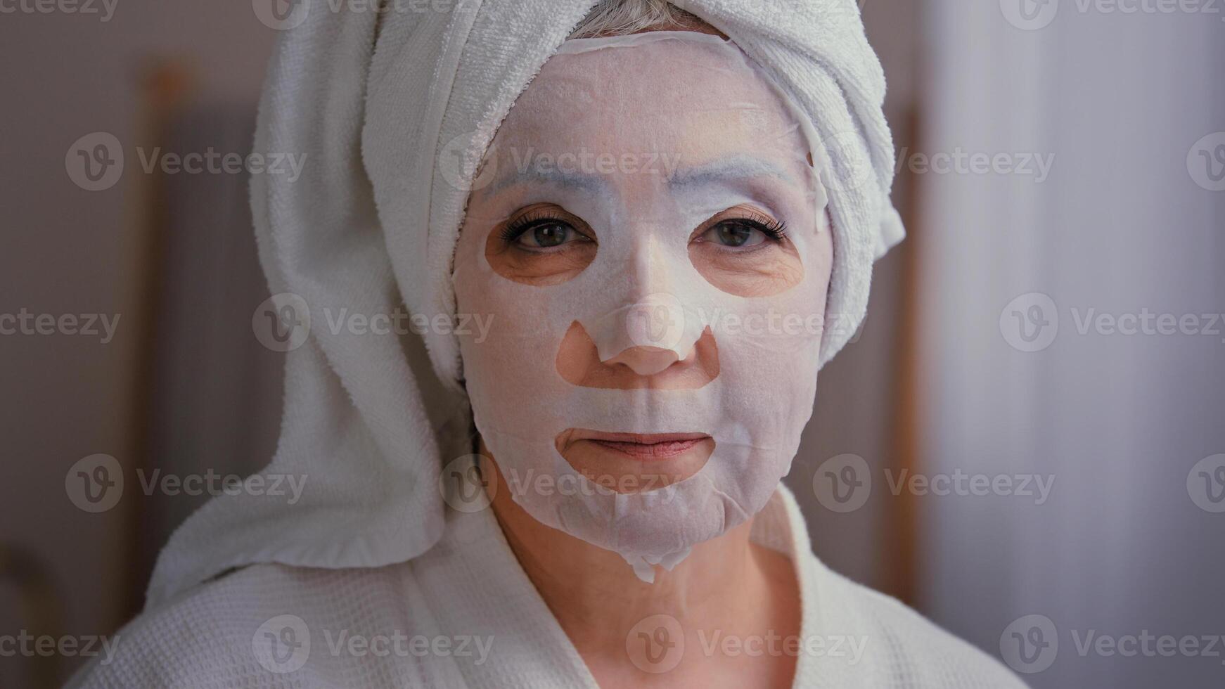 Old senior mature Caucasian woman apply skincare textile skin mask on female face showing ok good beauty cosmetology procedure in bathroom 60s lady wrap towel on head put moisturizing lifting cosmetic photo