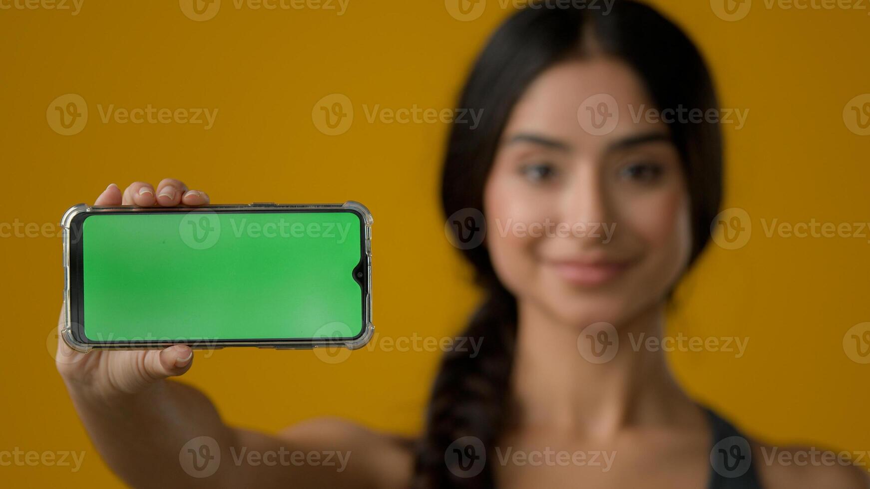 Close up Indian ethnic smiling woman female girl in yellow studio holding phone horizontal showing green empty mobile screen of smartphone chroma key display for advertisement online add offer in cell photo