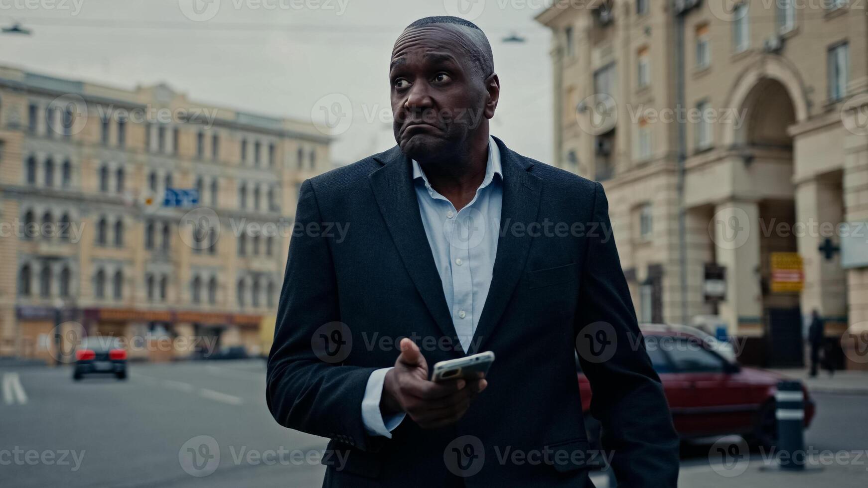 Ethnic adult man African middle-aged businessman entrepreneur with phone in city booking taxi with mobile app waiting car confused upset looking around problem with navigation mistake navigator wait photo