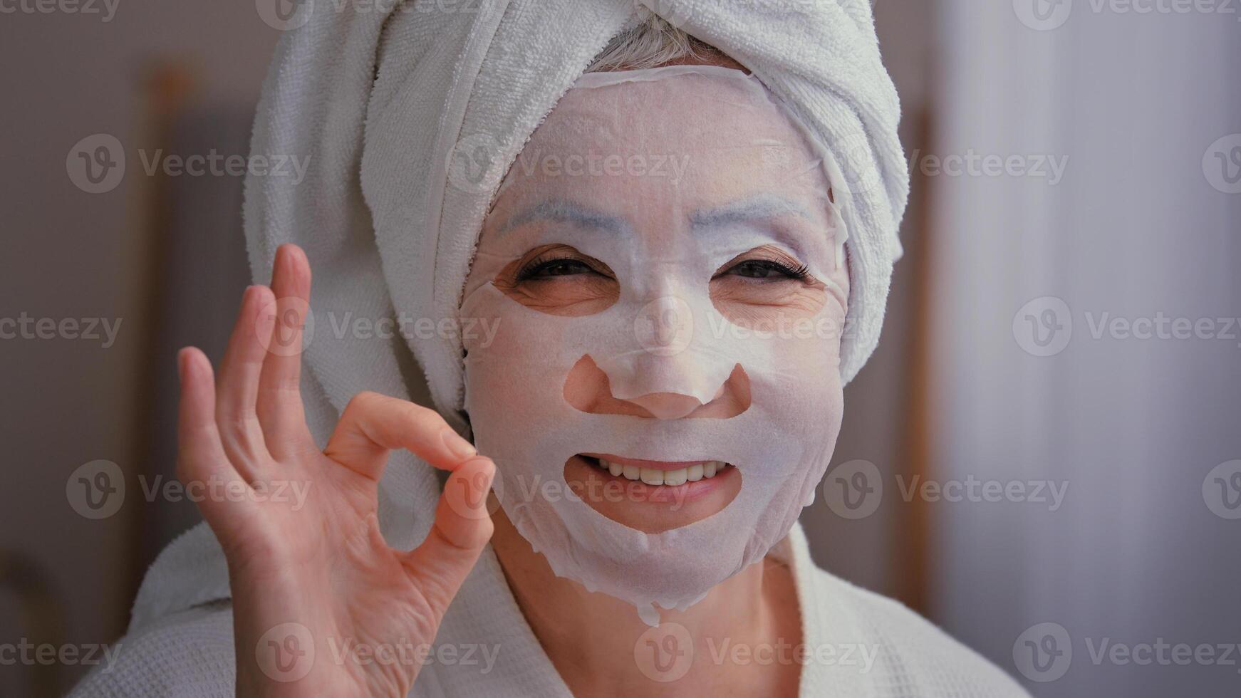 Close up old senior mature Caucasian woman with skincare textile skin mask on female face showing ok good beauty cosmetology procedure 60s lady wrap towel on head put moisturizing lifting cosmetic photo