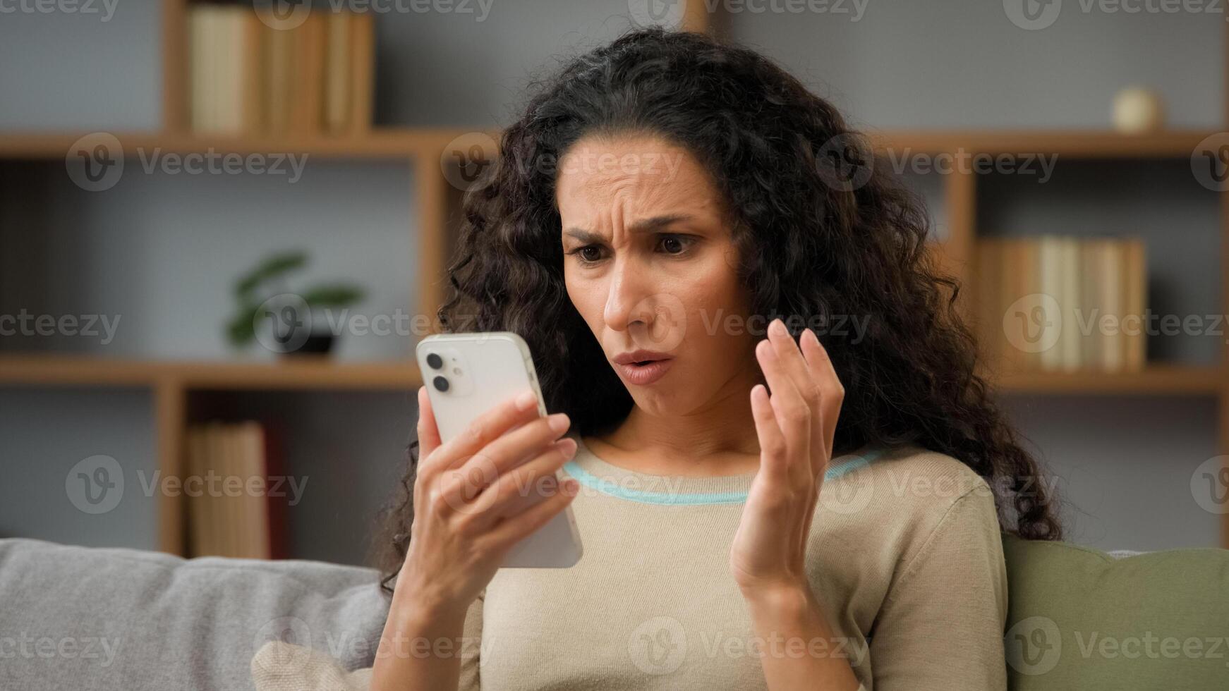Hispanic sad displeased woman has problem with online mobile app on cell phone unhappy with blocked account in social networks looking at cellphone screen scam unsuccessful bad news sitting at home photo