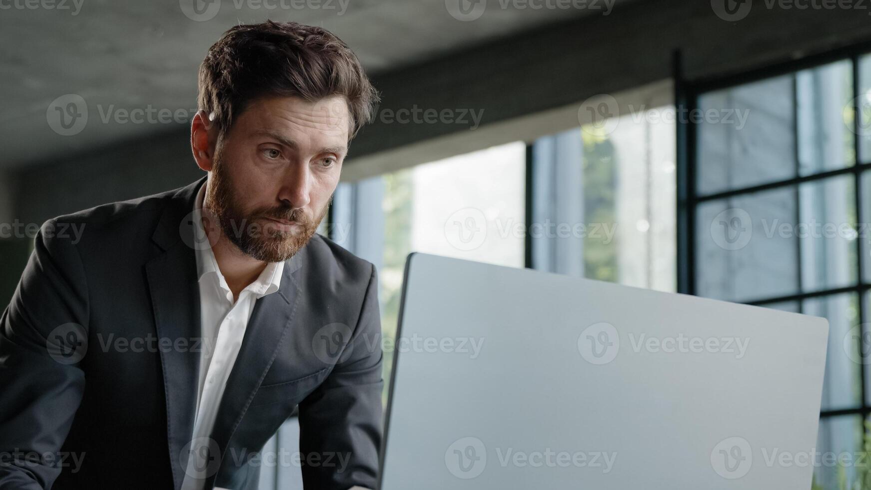 Serious 40s bearded businessman entrepreneur employee standing in office looking at papers report information papers check data in online net service in computer typing working paperwork deadline photo