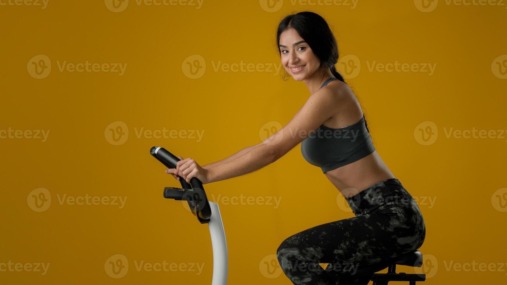 Indian ethnic strong slim sporty woman female lady athletic girl biking flexing cycling on exercise bike equipment riding sport fitness workout training healthy sports ride in yellow studio background photo
