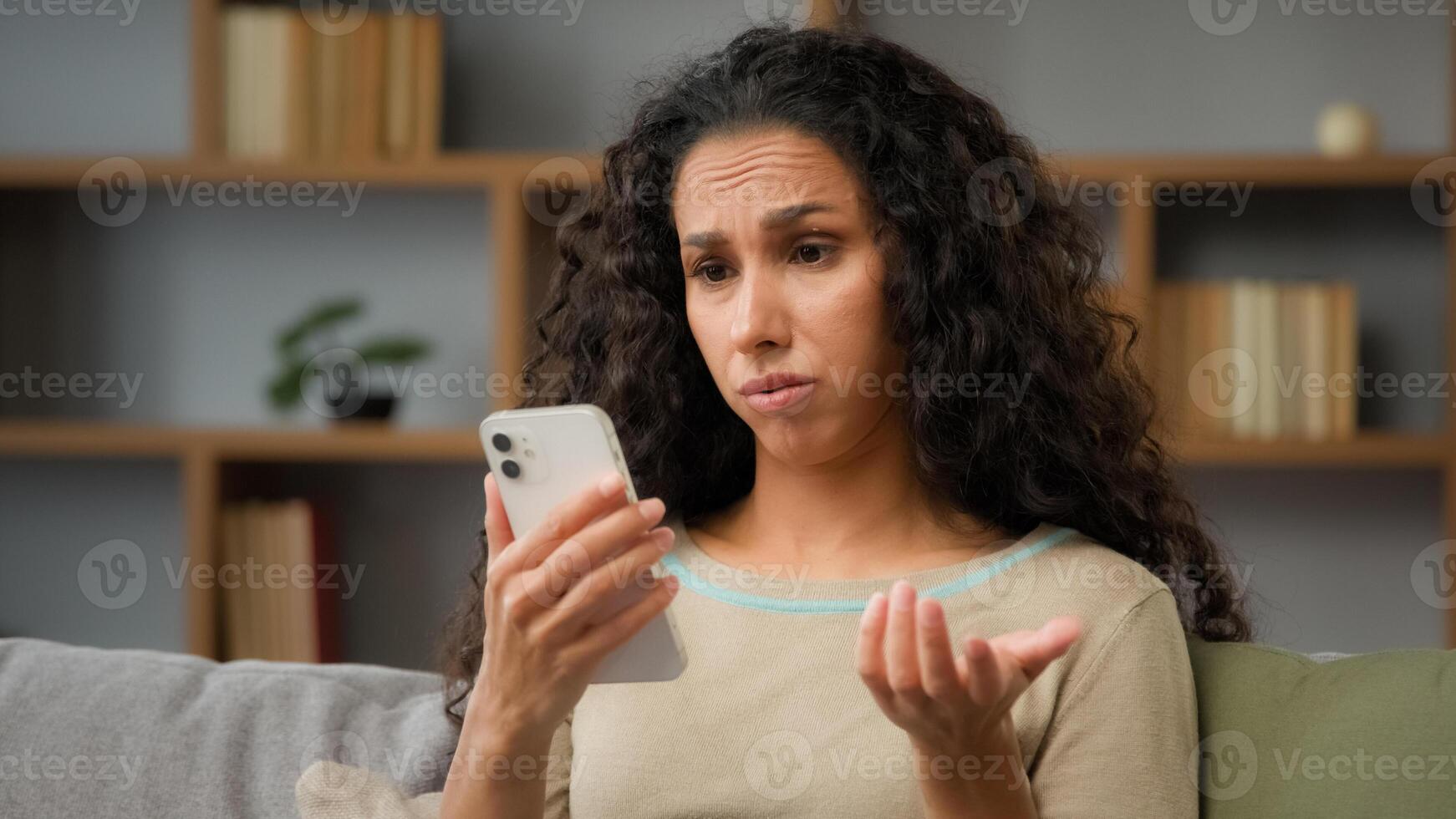 Hispanic sad displeased woman has problem with online mobile app on cell phone unhappy with blocked account in social networks looking at cellphone screen scam unsuccessful bad news sitting at home photo