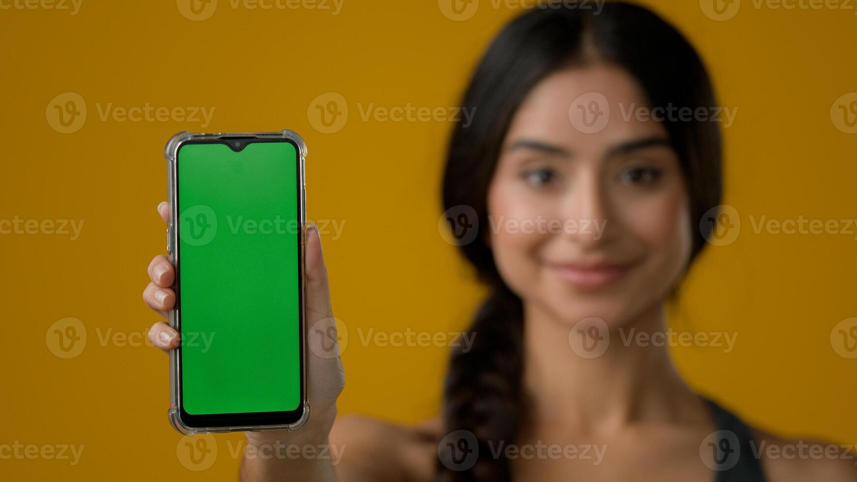 Indian ethnic woman lady girl lady in yellow studio background showing smartphone mobile telephone screen with green empty chroma key touchscreen technology cell message display advertise web service photo