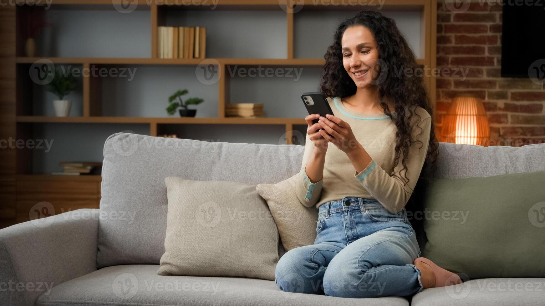 Smiling Hispanic Caucasian 30s woman housewife girl sitting on couch cozy sofa at home using smartphone for ordering food delivery chatting with mobile app looking at phone screen with smile browsing photo