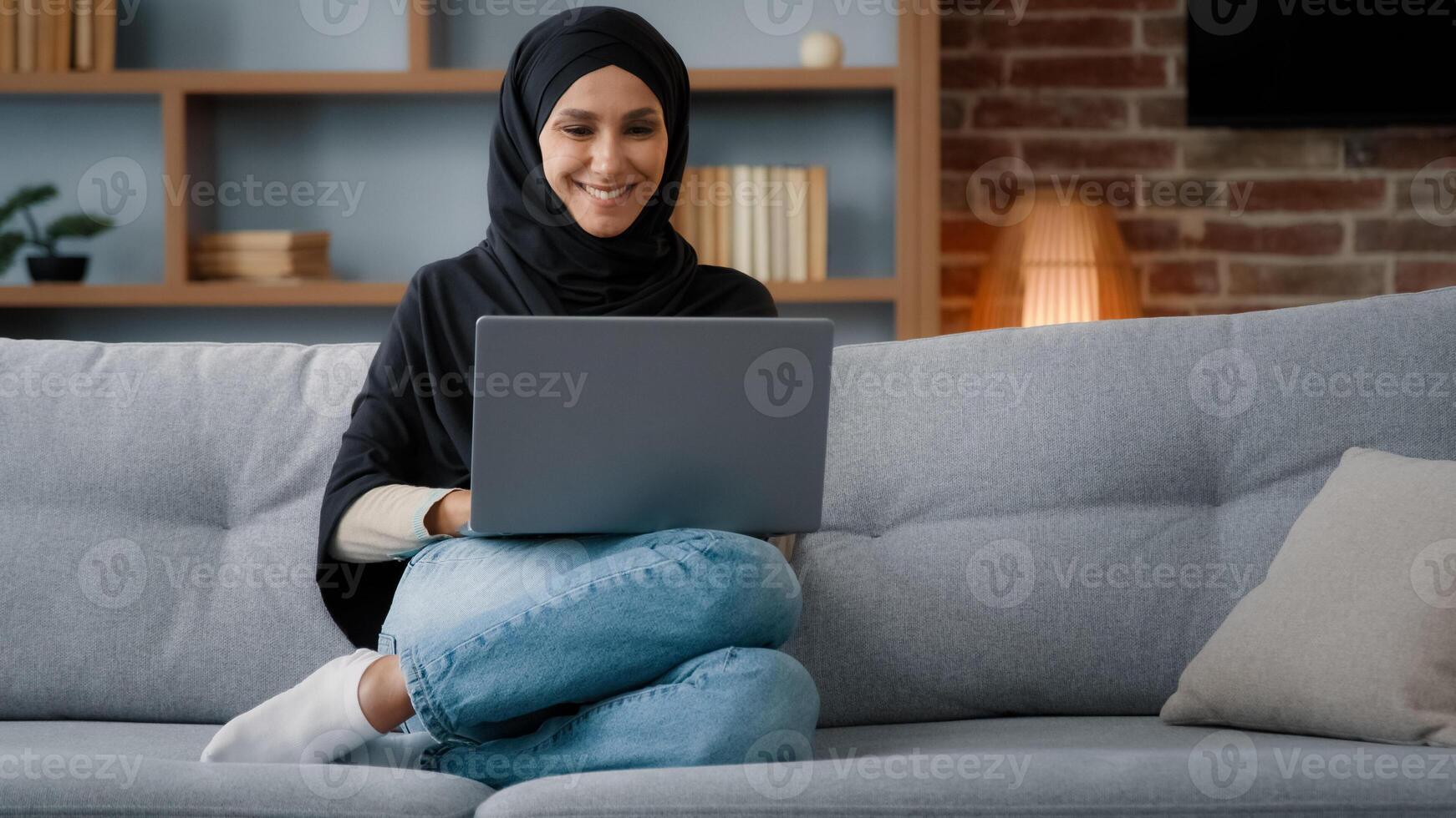 Muslim Islamic woman in hijab sitting at home living room relaxing on sofa couch using laptop online website ordering booking in internet shop working studying remote distant chatting typing computer photo
