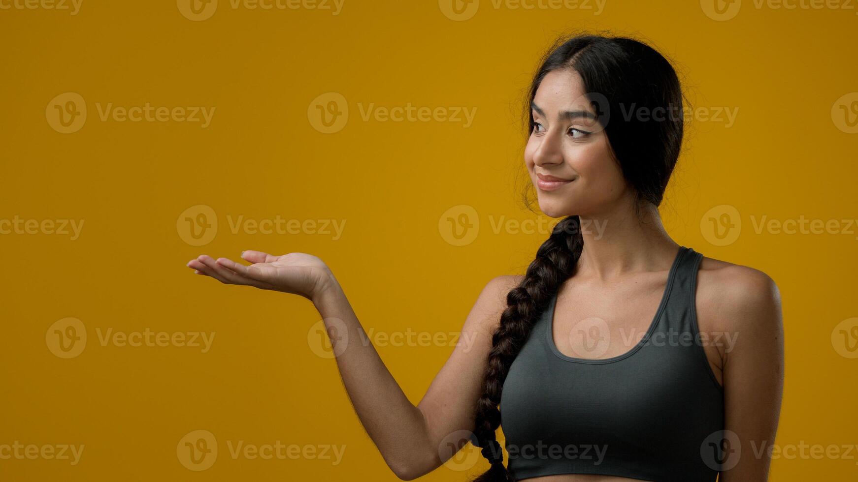Portrait Indian 20s woman in studio yellow background pointing to side at palm arm hand point advertisement aside, promotional recommend recommendation prize sign girl lady showing to empty cope space photo