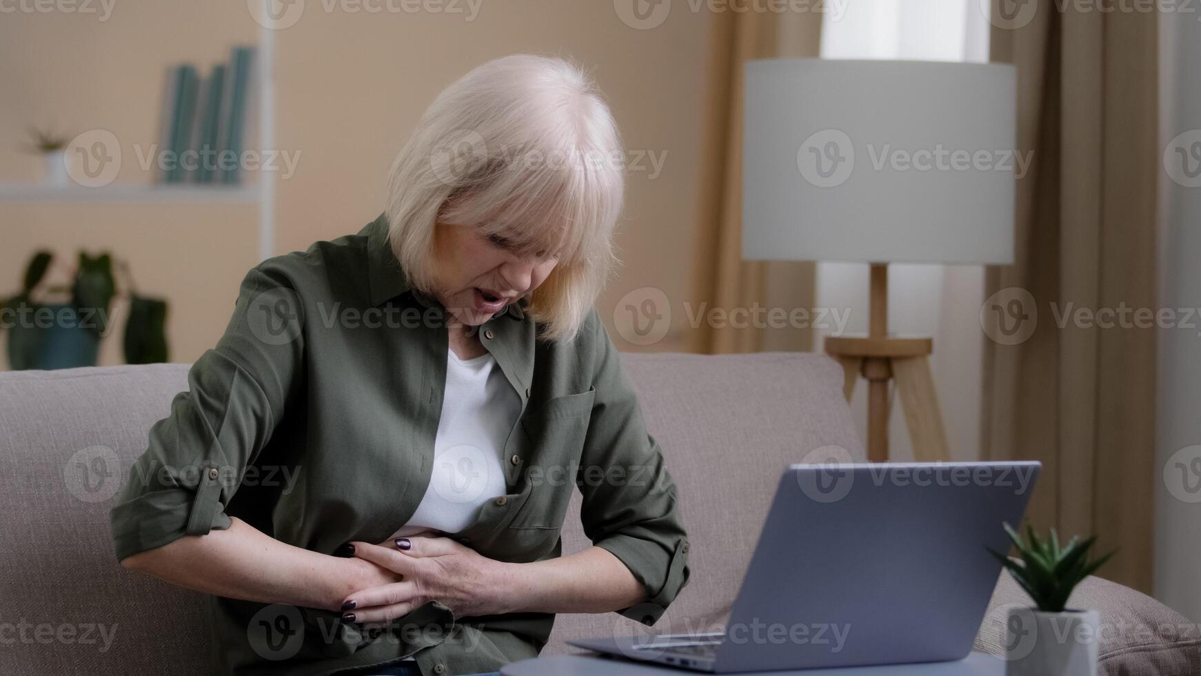 Sick 50s woman with laptop bad pain hold belly stomach suffer with hurt abdomen ache at home, old lady feels gastritis pancreatitis symptom sit on couch, upset senior with indigestion flatulence photo