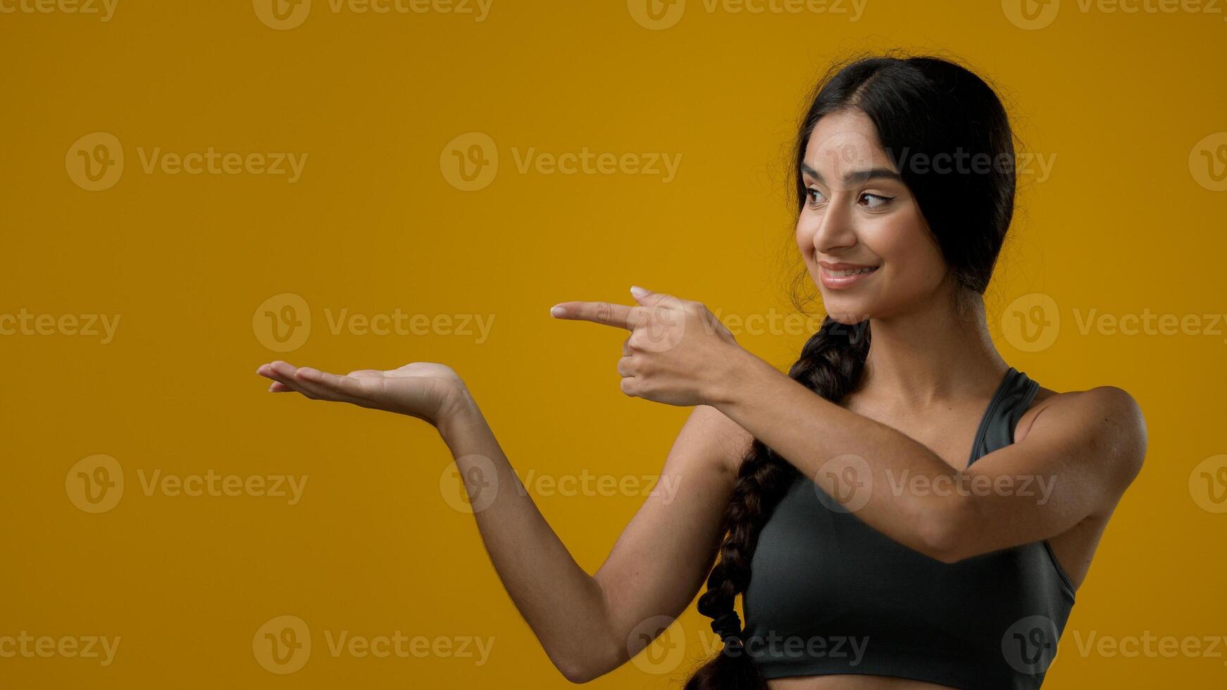 Portrait in studio Indian 20s woman posing on yellow background pointing to side at palm arm hand point advertisement aside recommend recommendation prize sign girl lady showing to empty copy space photo